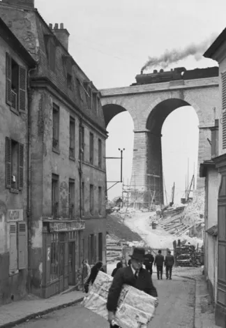 Photo noir et blanc d'une rue urbaine bordée d'immeubles anciens, avec un train à vapeur franchissant un viaduc en arches à l'arrière-plan et des passants au premier plan.