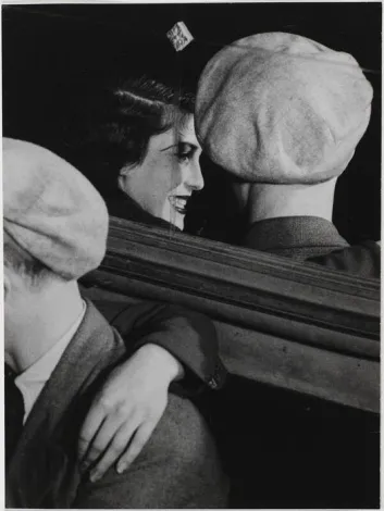 Photo noir et blanc d'une jeune femme souriante, entourée de deux hommes coiffés de casquettes plates, dans une scène de proximité et de convivialité.