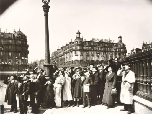 Photo noir et blanc d'une foule parisienne regardant le ciel, la main en visière, sur un pont urbain bordé d'immeubles haussmanniens, probablement lors d'une éclipse solaire.