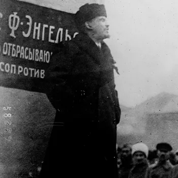 Photo noir et blanc de Lénine, en manteau et chapka, debout sur une estrade et haranguant une foule, à côté d'une pancarte portant du texte russe mentionnant Engels.