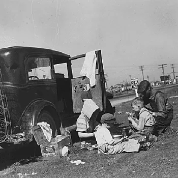 Photo noir et blanc d'une famille de migrants installée au bord d'une route près de leur voiture chargée d'affaires, pendant la Grande Dépression.