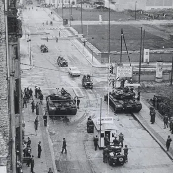Photo noir et blanc de chars et de soldats face à face de part et d'autre d'un poste de contrôle militaire, avec un panneau frontalier américain visible — scène évoquant le point de passage de Checkpoint Charlie à Berlin.