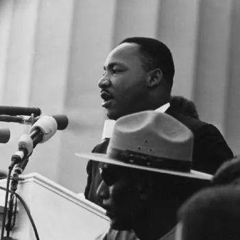 Photo noir et blanc de Martin Luther King prononçant un discours au micro, avec un garde en uniforme et chapeau au premier plan — image associée à la marche sur Washington de 1963.