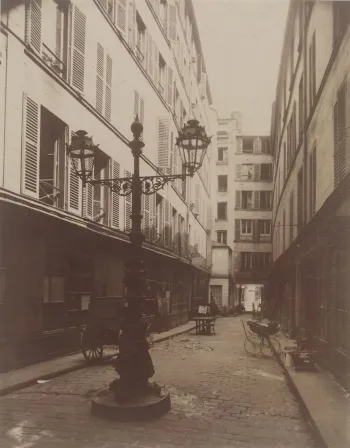 Photo sépia d'une cour intérieure parisienne pavée, avec un réverbère ouvragé, une voiture d'enfant ancienne et des façades aux volets, par Eugène Atget.